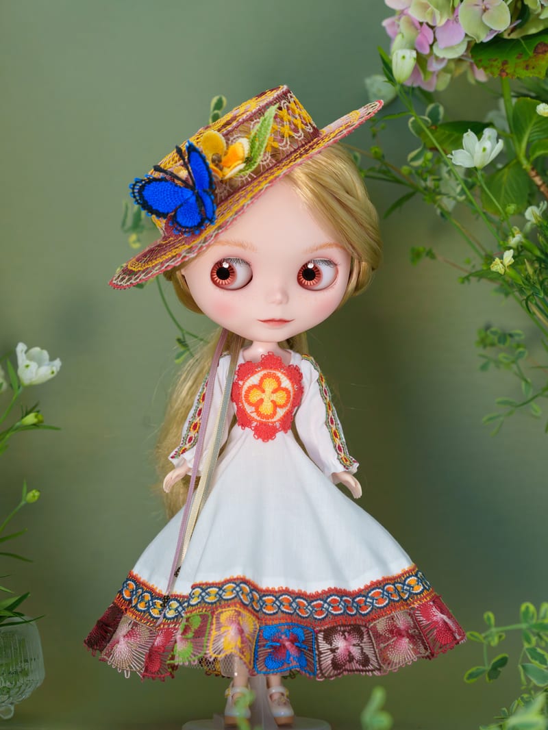 A Blythe doll in a white dress with Ñandutí lace trim and a lace hat, standing among hydrangea.