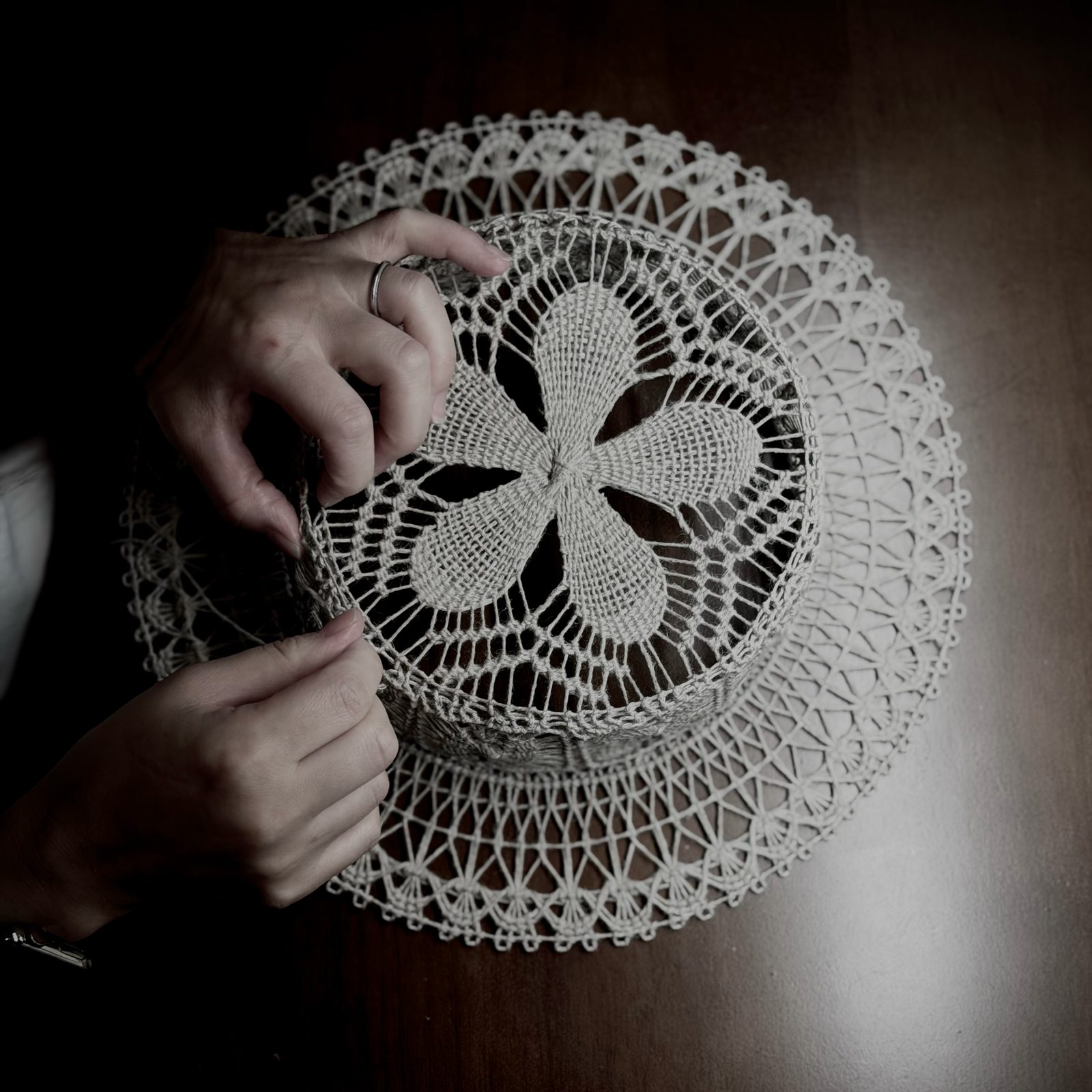 Two hands working a ramie Ñandutí hat on a wooden table, its crown worked outward from the centre.